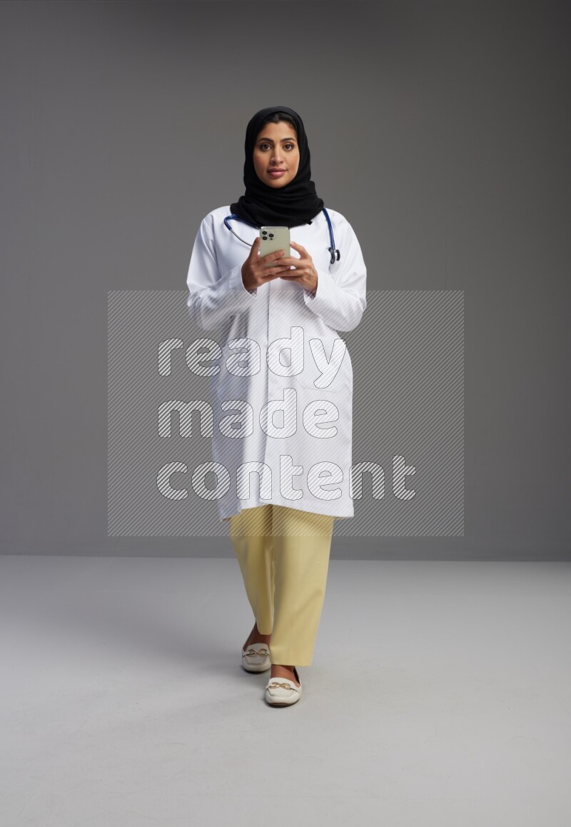 Saudi woman wearing lab coat with stethoscope standing texting on phone on Gray background