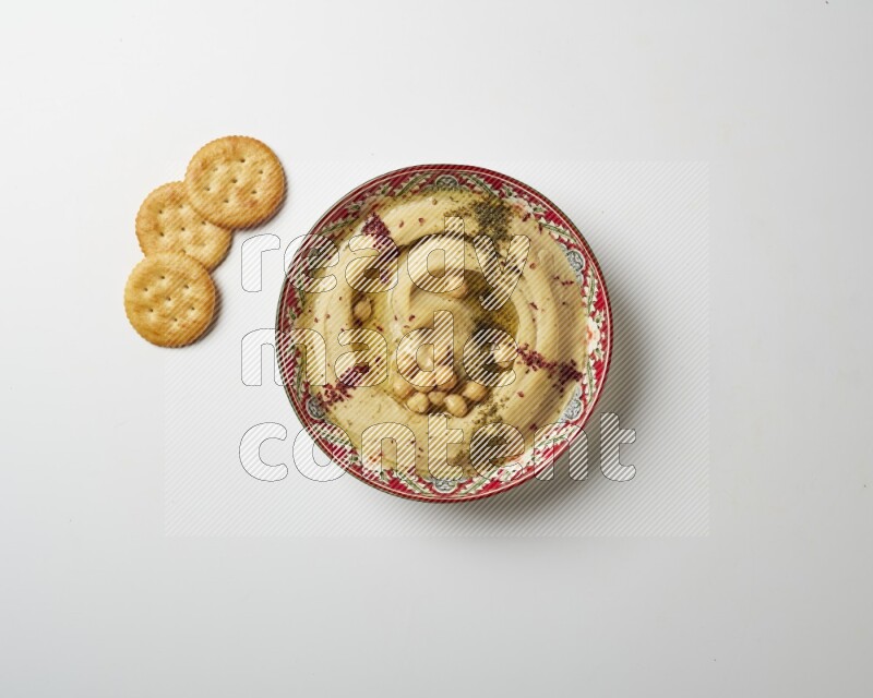 Hummus in a red plate with patterns garnished with zaatar & sumak on a white background