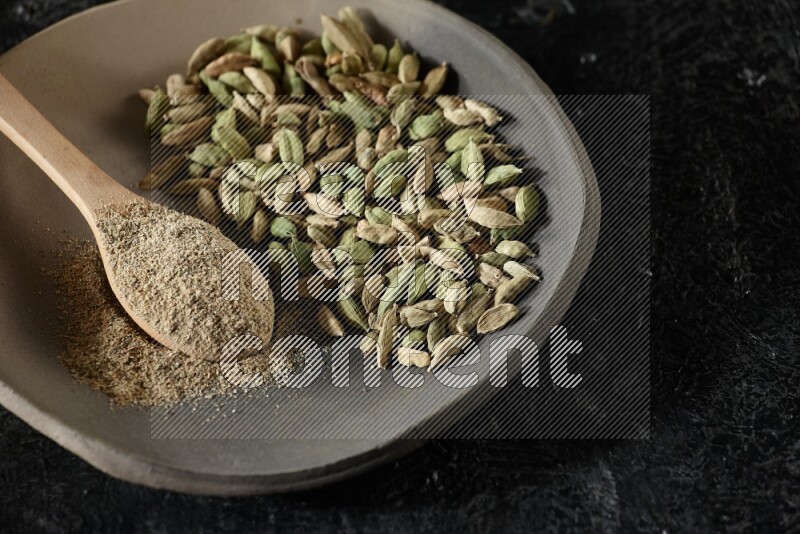 A plate filled with cardamom seeds and a wooden spoon full of cardamom powder on a textured black flooring