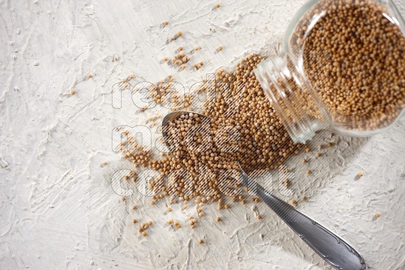 A glass spice jar and a metal spoon full of mustard seeds and jar is flipped with fallen seeds on a textured white flooring