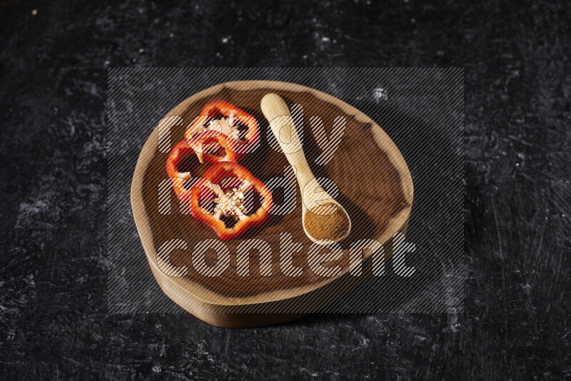 A wooden spoon full of ground paprika powder with red bell pepper slices beside it, all on a wooden tray on black background