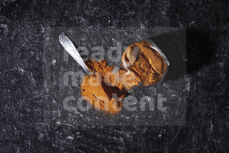 A glass jar full of ground paprika powder flipped with some spilling powder on black background