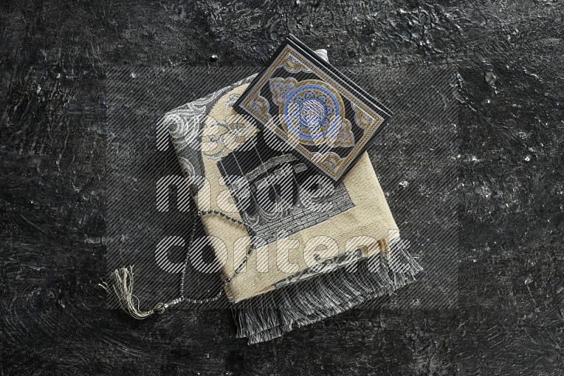 A folded prayer mat with different elements such as quran and prayer beads on black textured background