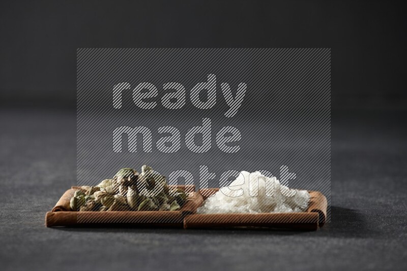 مربعان من أعواد القرفة ممتلئان بالملح الأبيض وحبوب الهيل علي خلفية سوداء