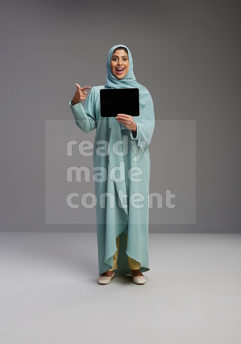 Saudi Woman wearing Abaya standing showing tablet to camera on Gray background