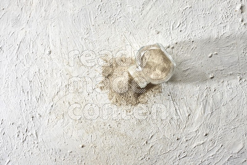 A flipped herbal glass jar full of white pepper powder with spilled powder on textured white flooring