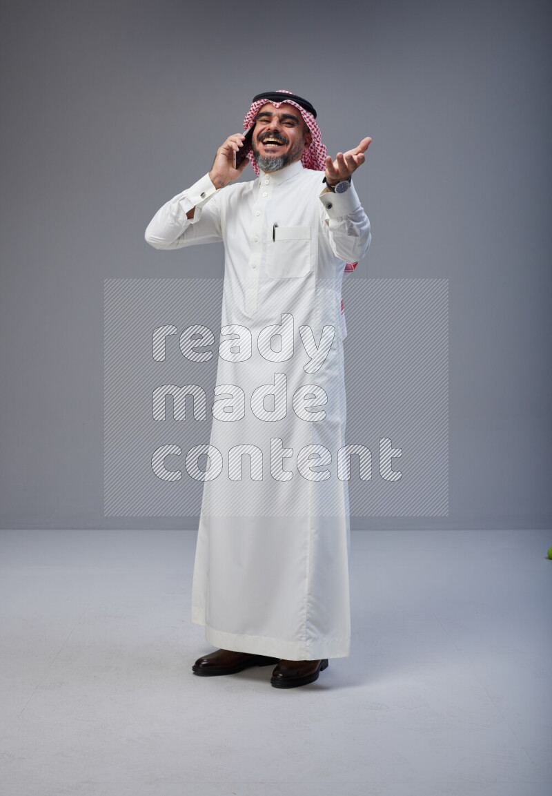 Saudi man Wearing Thob and red Shomag standing talking on phone on Gray background