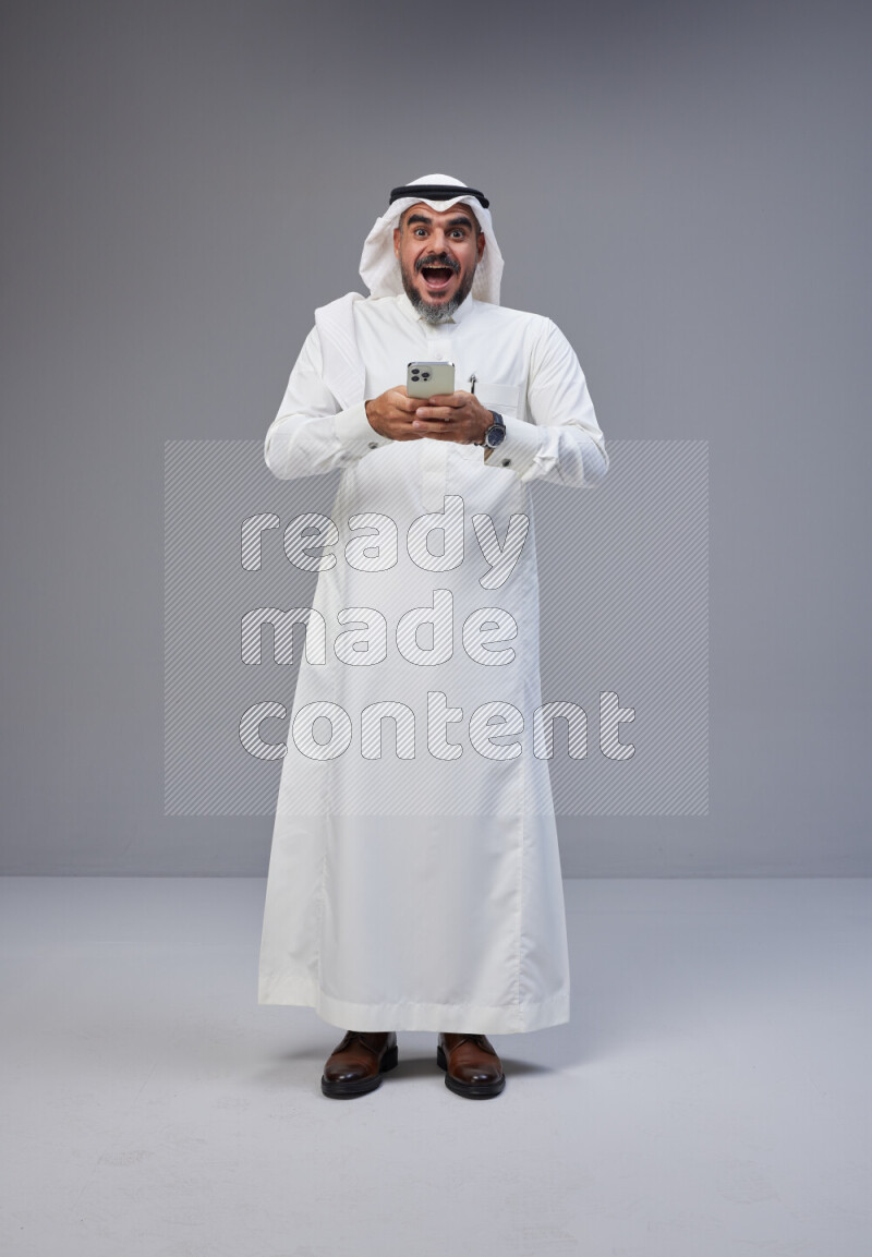 Saudi man Wearing Thob and white Shomag standing texting on phone on Gray background