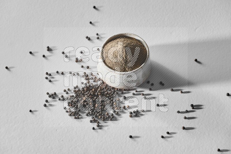 فلفل أسود مطحون في وعاء أبيض من السيراميك مع حبوب فلفل أسود علي خلفية بيضاء