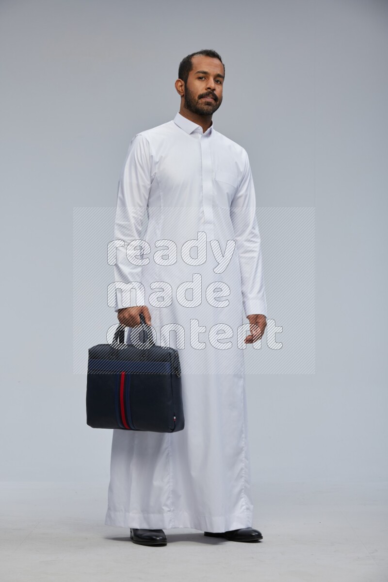 Saudi man wearing thob standing holding suitcase on gray background