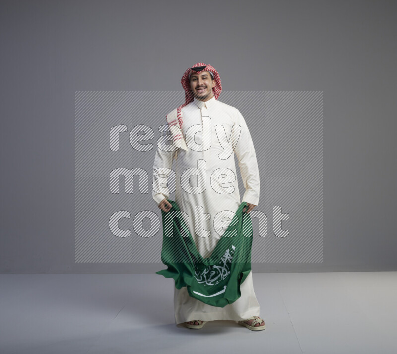 A saudi man standing wearing thob and red shomag holding big saudi flag on gray background
