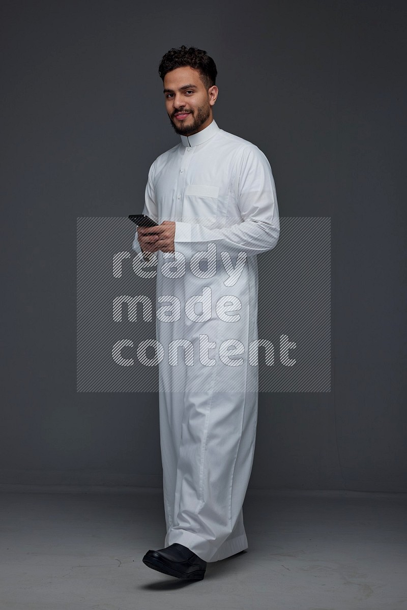 A Saudi man wearing Thobe standing and using his phone eye level on a gray background
