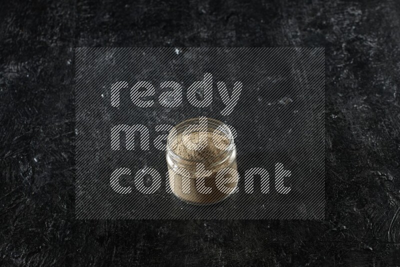 A glass jar full of cardamom powder on textured black flooring