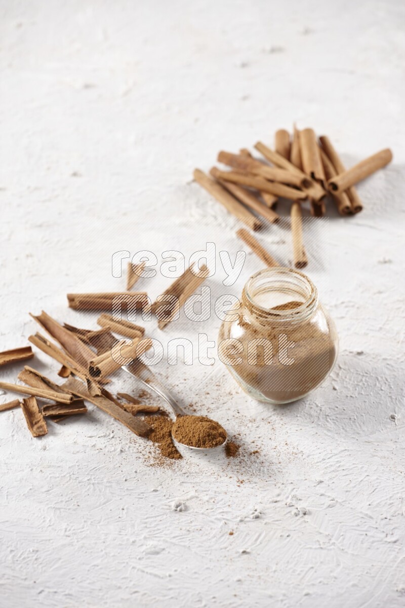 Herbal glass jar full cinnamon powder and a metal spoon surrounded by cinnamon sticks on a white background