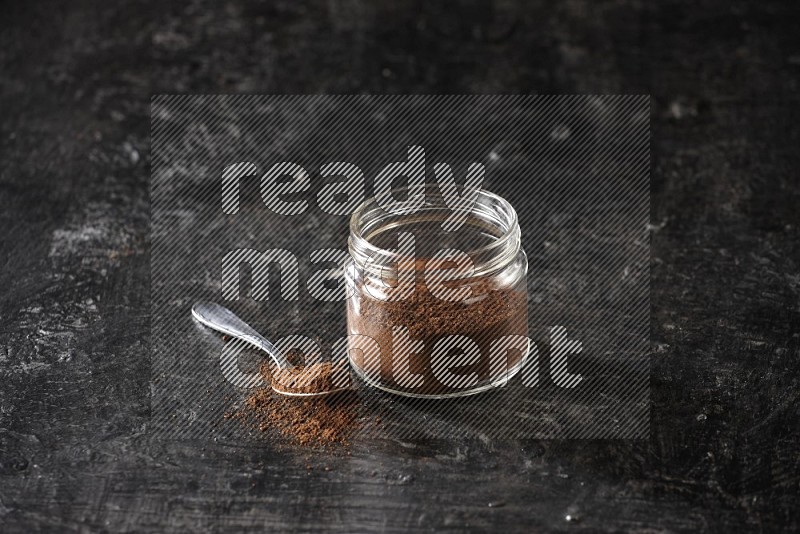 A glass jar full of cloves powder with a metal spoon on a textured black flooring