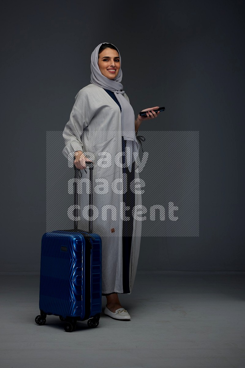 A Saudi woman wearing a light gray Abaya and head scarf standing holding a phone and being shocked  eye level on a grey background