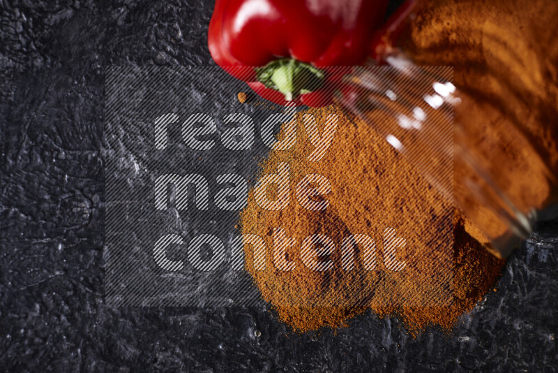 A glass jar full of ground paprika powder flipped with some spilling powder on black background