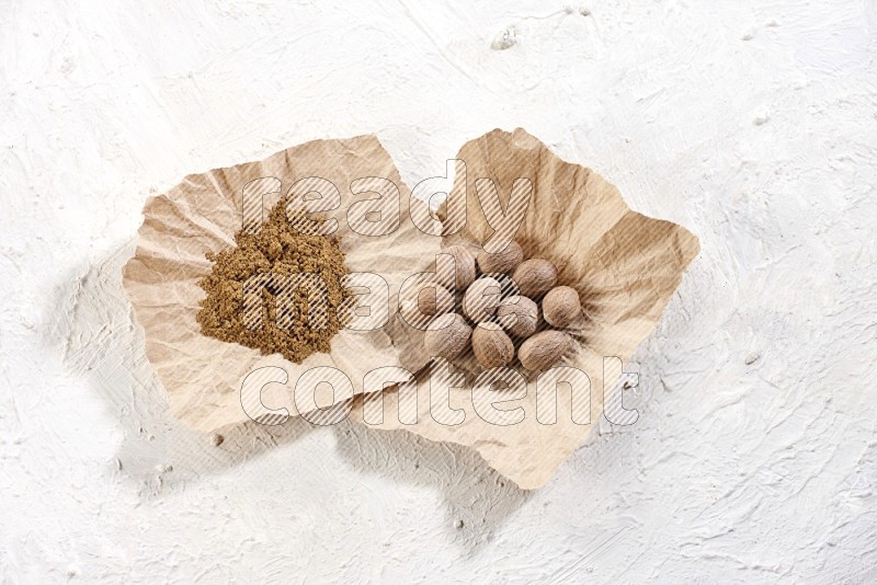 2 crumpled piece of paper full of nutmeg seeds and powder on a textured white flooring in different angles