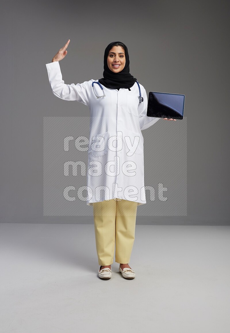 Saudi woman wearing lab coat with stethoscope standing showing tablet to camera with sign in the back on Gray background