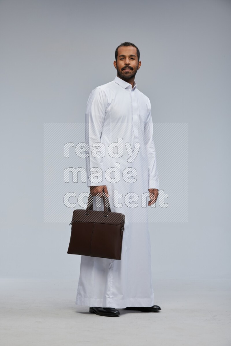 Saudi man wearing thob standing holding suitcase on gray background