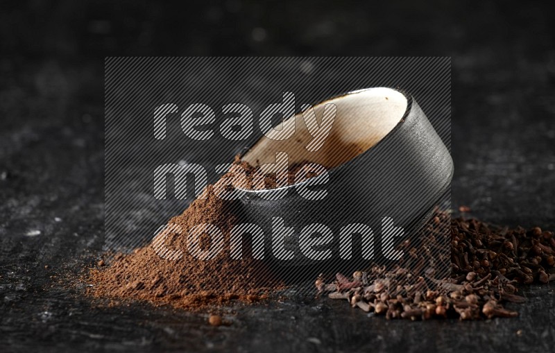 A black pottery bowl full of cloves powder spilled out of it with whole grains on a textured black flooring