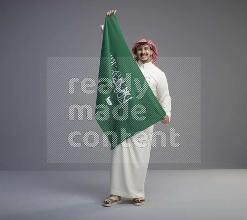 A saudi man standing wearing thob and red shomag holding big saudi flag on gray background
