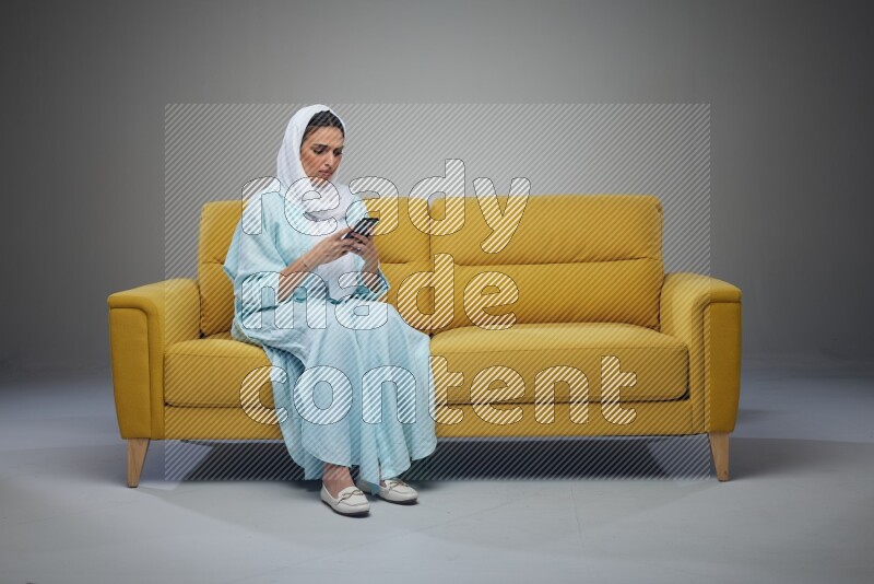 A Saudi woman wearing a light blue Abaya and white head scarf sitting on a yellow sofa and using her phone eye level on a grey background