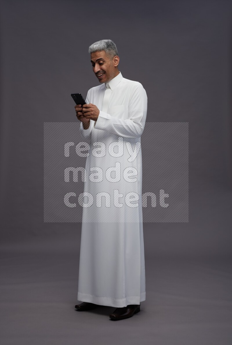 Saudi man wearing thob standing texting on phone on gray background