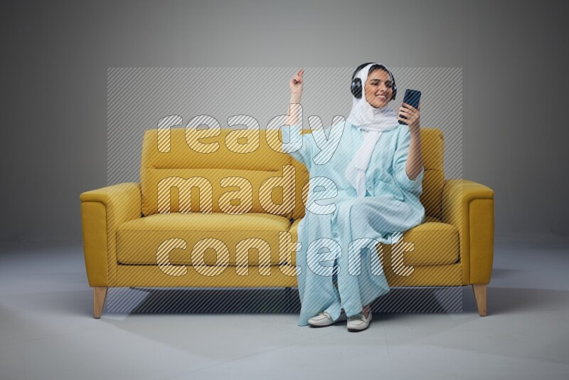 A Saudi woman wearing a light blue Abaya and a white head scarf sitting on a yellow sofa and holding an iPad while wearing headphone eye level on a grey background