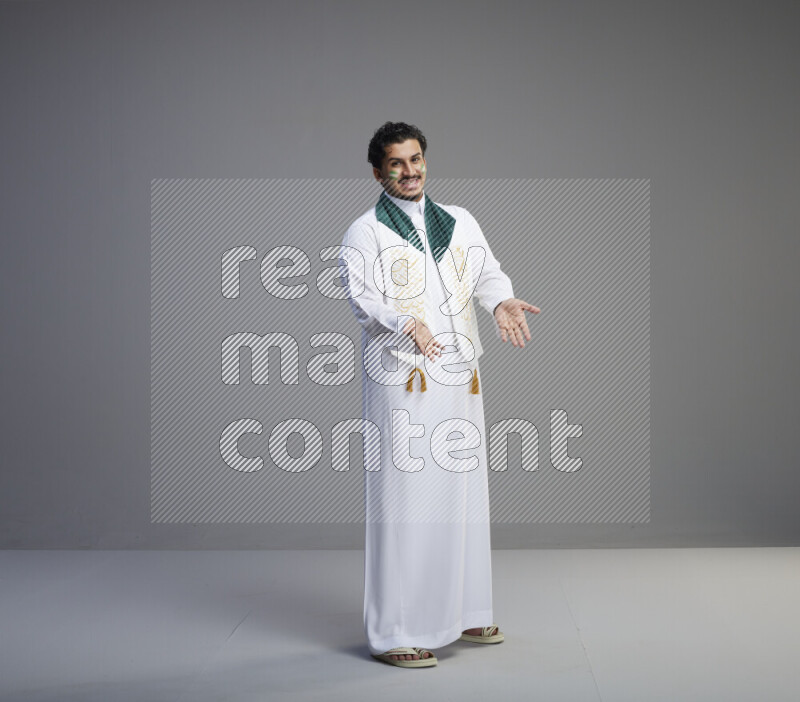 A Saudi man standing wearing thob and Saudi flag scarf with face painting on gray background