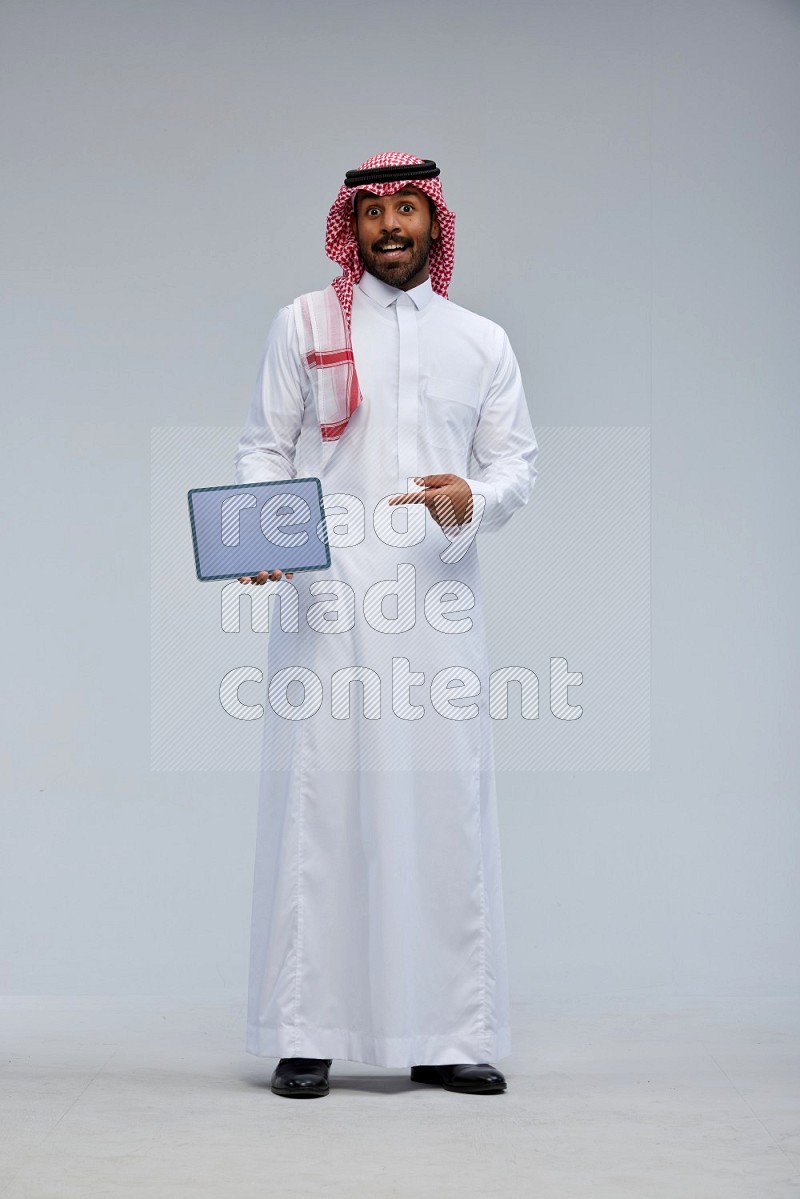 Saudi man Wearing Thob and shomag standing showing tablet to camera on Gray background