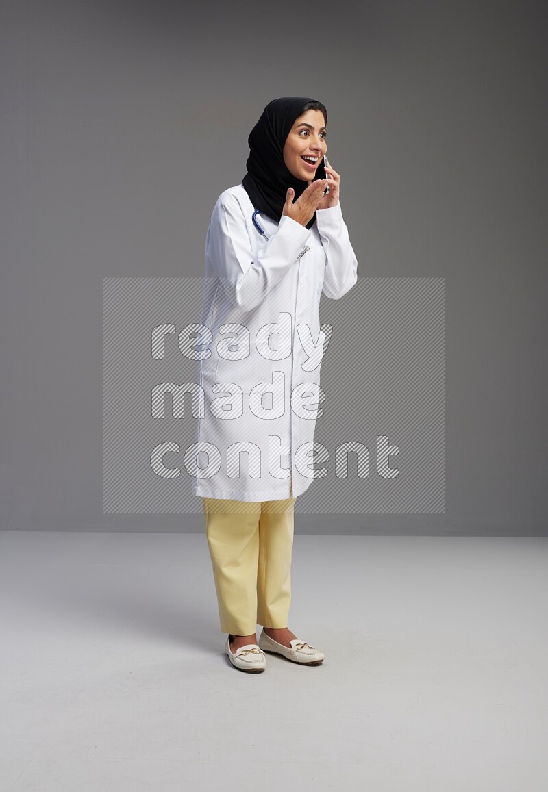 Saudi woman wearing lab coat with stethoscope standing talking on phone on Gray background