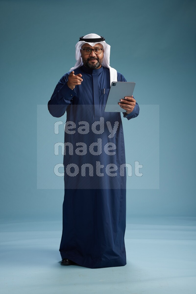 Saudi Man with shimag Standing holding ATM card while working on tablet on blue background