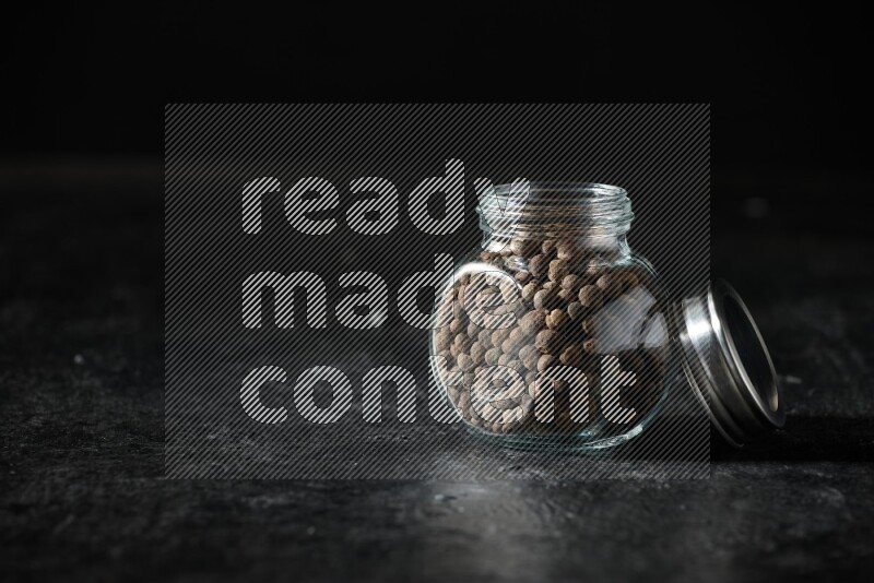 A glass spice jar full of allspice whole balls on a textured black flooring