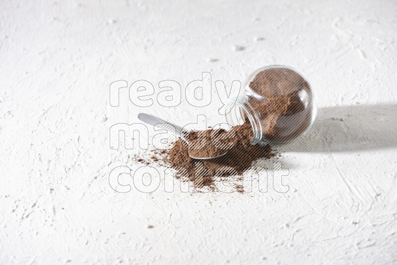 A flipped glass spice jar and a metal spoon full of cloves powder and powder came out of the jar on textured white flooring