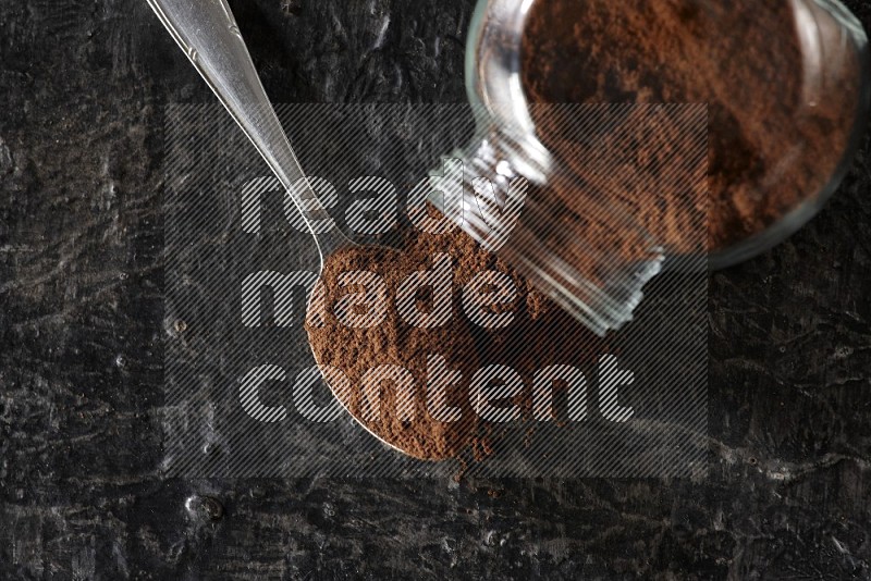 A flipped glass spice jar and a metal spoon full of cloves powder on textured black flooring