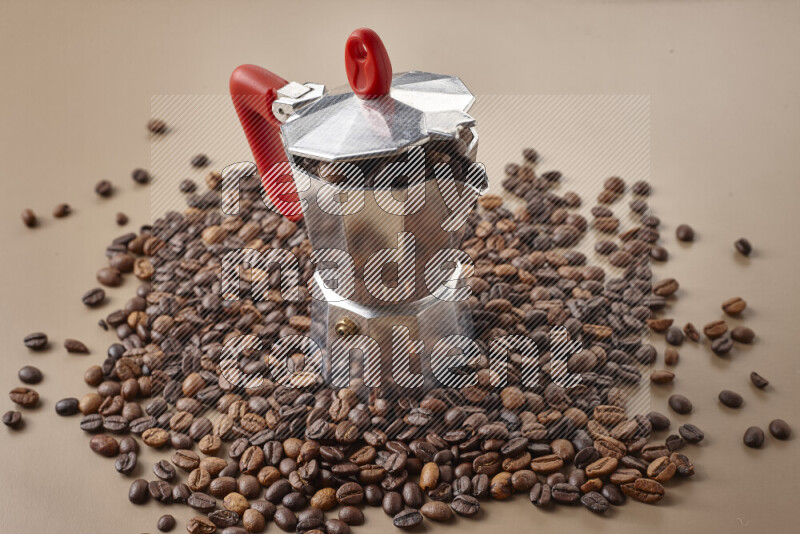 A moka pot with red handle surrounded by roasted coffee beans on beige background