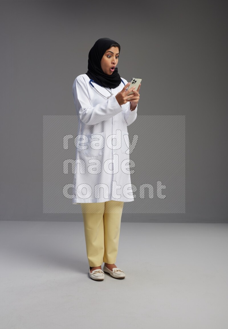 Saudi woman wearing lab coat with stethoscope standing texting on phone on Gray background