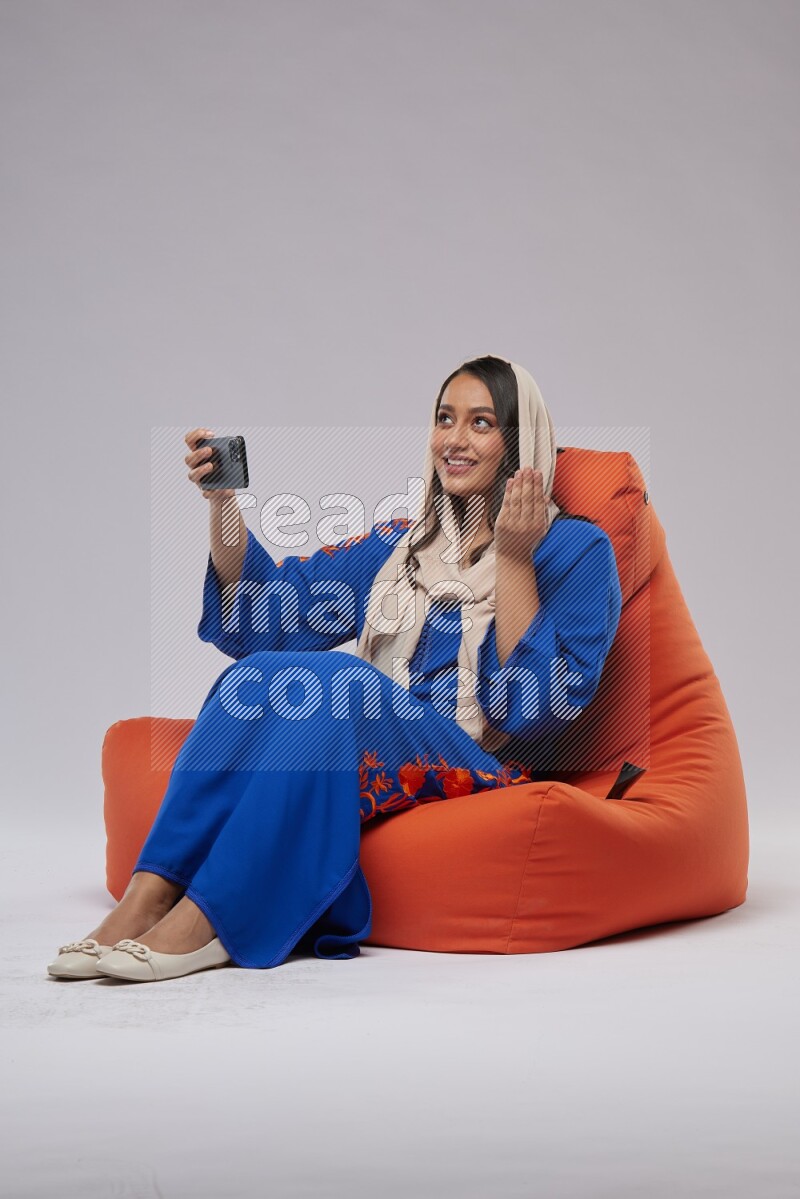 A Saudi woman wearing jalabiya sitting on an orange beanbag and taking selfie