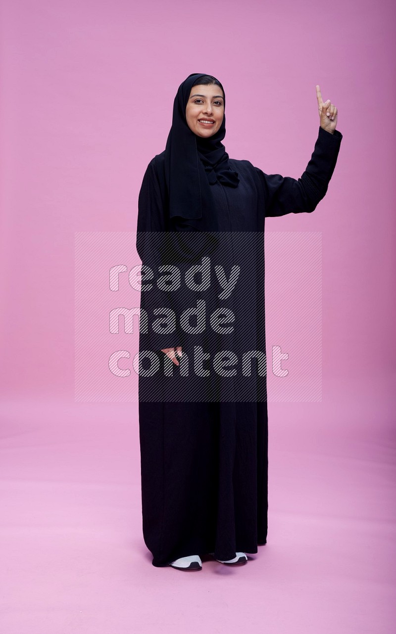 Saudi woman wearing Abaya standing interacting with the camera on pink background