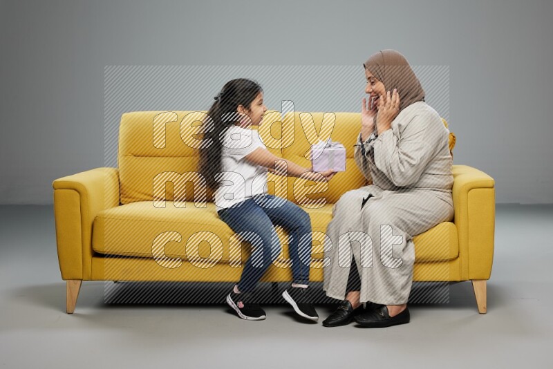 A girl sitting on a yellow sofa and giving a gift to her mother on gray background