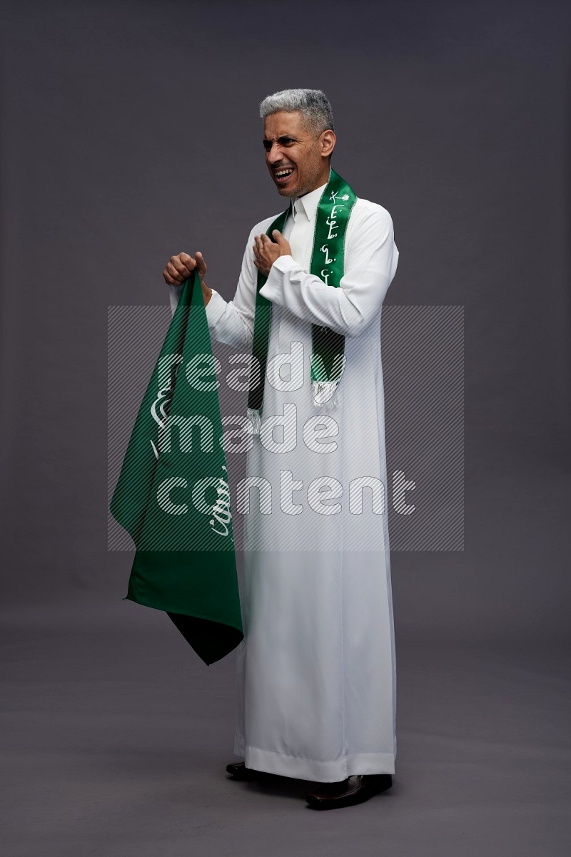 Saudi man wearing thob standing holding Saudi flag on gray background