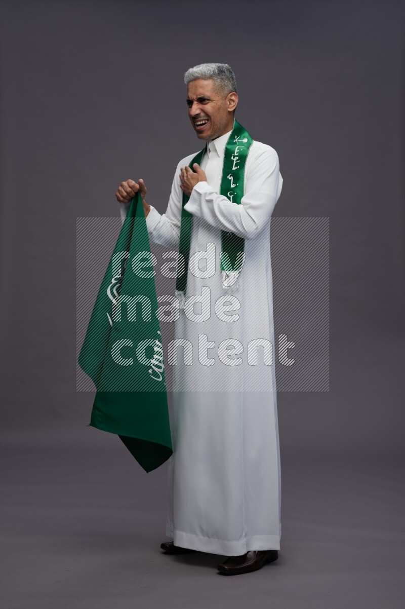 Saudi man wearing thob standing holding Saudi flag on gray background
