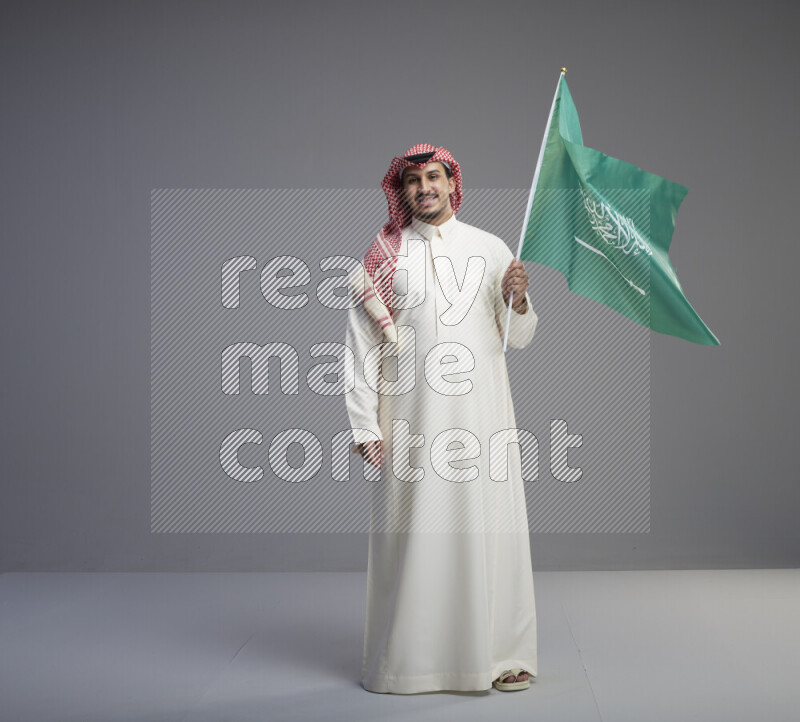A Saudi man standing wearing thob and red shomag raising big Saudi flag on gray background