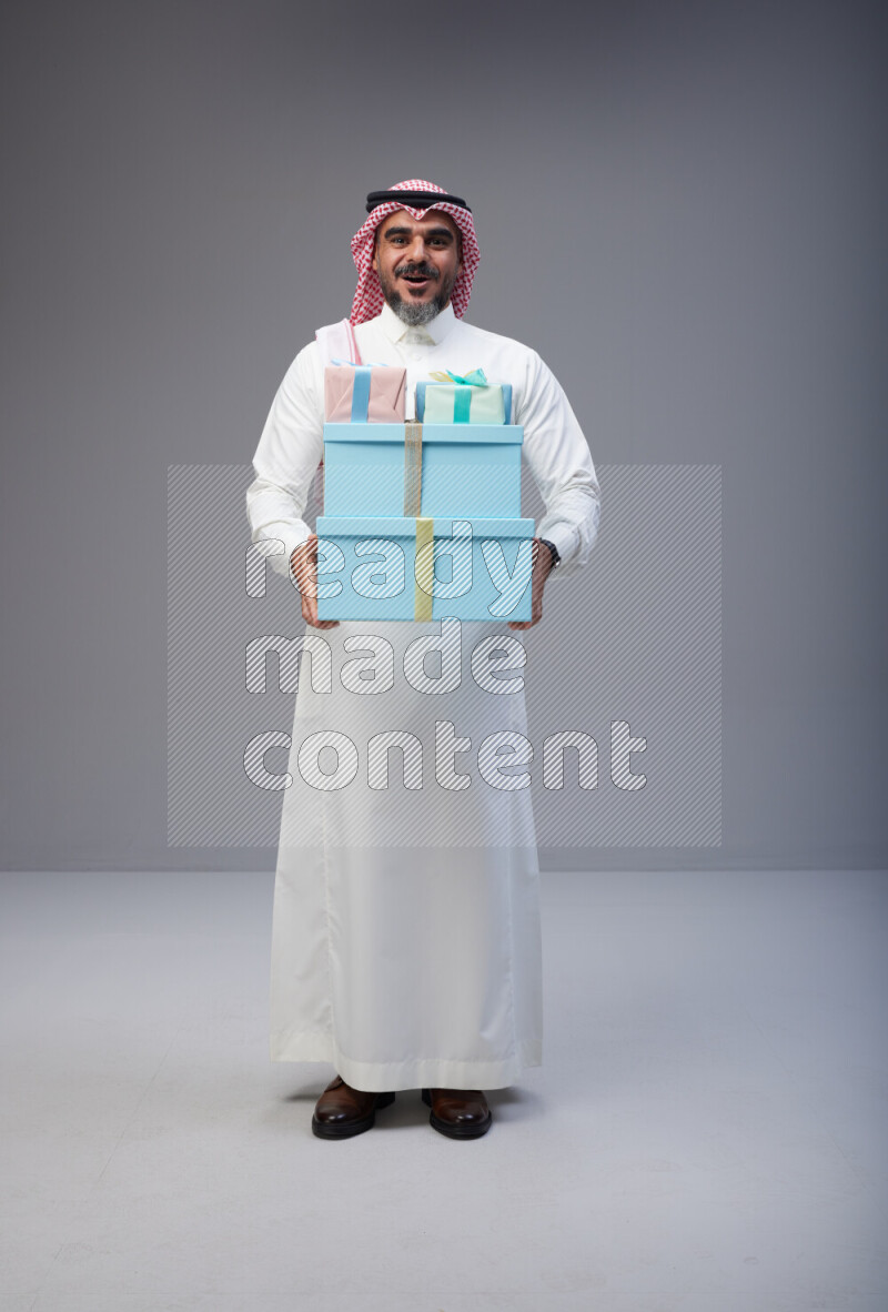 Saudi man Wearing Thob and red Shomag standing holding gift box on Gray background