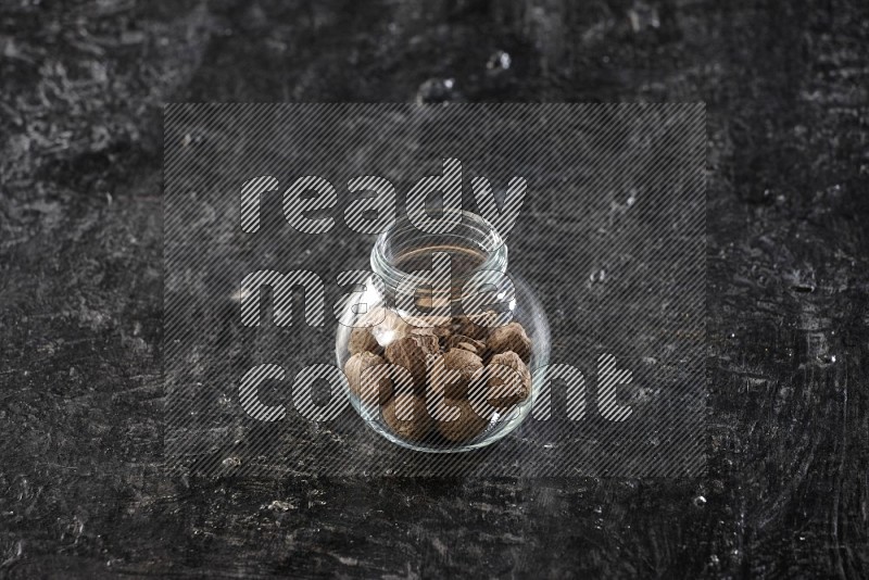 A glass spice jar full of nutmeg on a textured black flooring in different angles