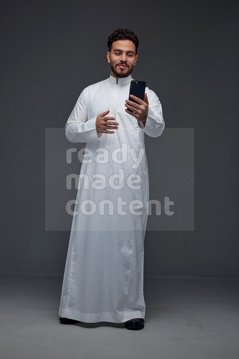 A Saudi man wearing Thobe and making a video call using his phone while standing and making different poses eye level on a gray background