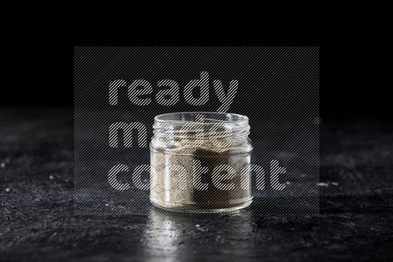 A glass jar full of white pepper powder on textured black flooring