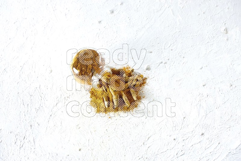 A flipped glass spice jar full of turmeric powder and powder spilled out of it with dried whole fingers on textured white flooring