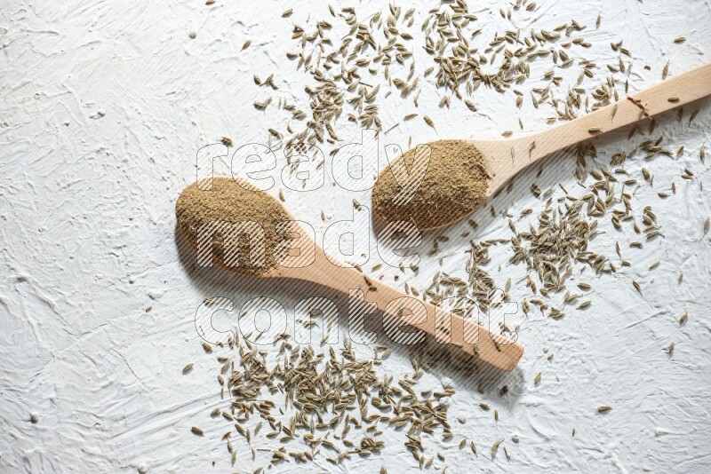 2 Wooden spoons full of cumin powder and cumin seeds on textured white flooring